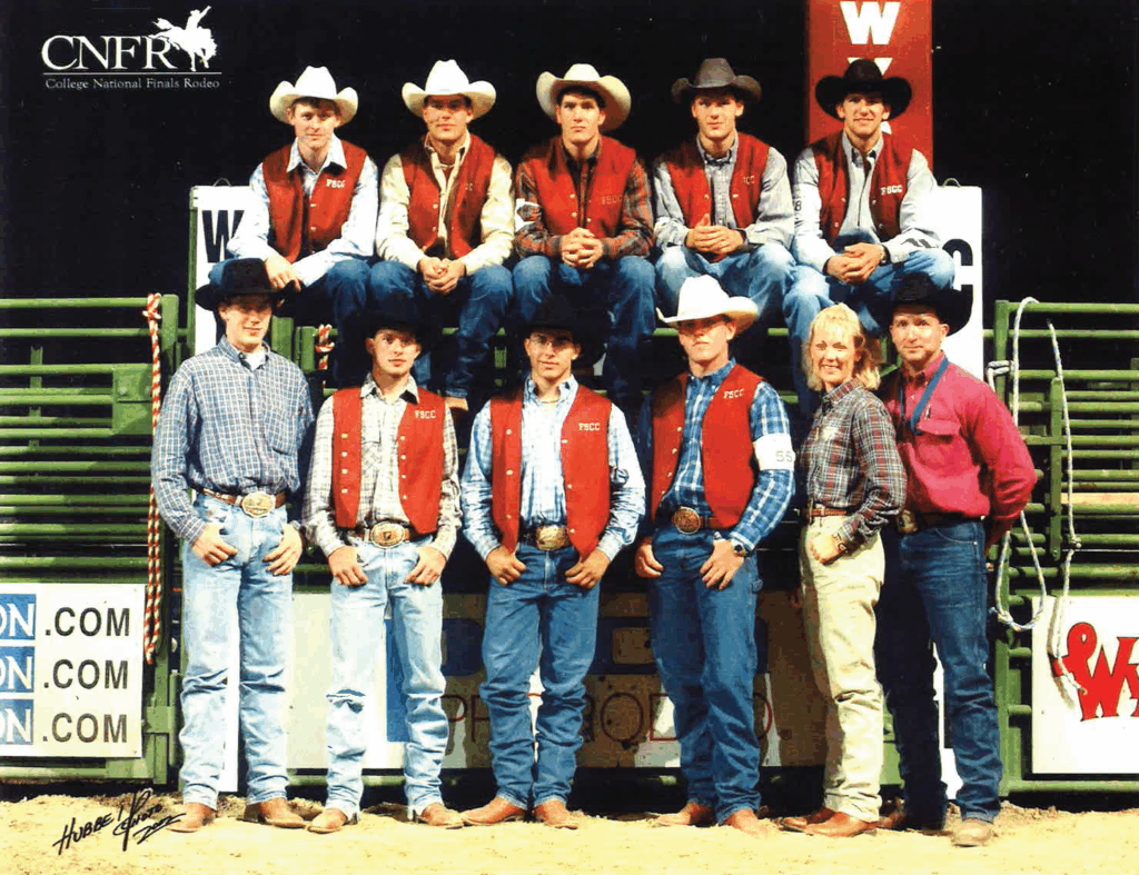 Team photo of the Fort Scott Community College rodeo members at the College National Finals Rodeo. Five young men, all in cowboy hats, FSCC vests, and denim, are seated on green chutes labeled 'WYO' with a CNFR sign above. Everyone is dressed in western attire, including boots, jeans, button-down shirts, and large belt buckles. The CNFR College National Finals Rodeo logo is at the top left. Green livestock fencing and arena advertisements are visible in the background, and the photo is set on a dirt arena floor.