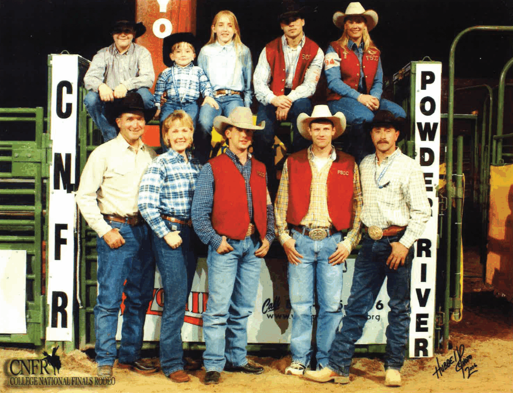 Group photo of the Fort Scott Community College rodeo team at the College National Finals Rodeo. Five team members are seated on and in front of a green and yellow rodeo chute labeled 'Powder River' and 'CNFR.' The group is dressed in western shirts, jeans, boots, and cowboy hats, with some wearing FSCC vests and belt buckles. The CNFR College National Finals Rodeo logo appears in the bottom left corner. The setting is an indoor arena with green livestock panels and an arena floor of dirt.