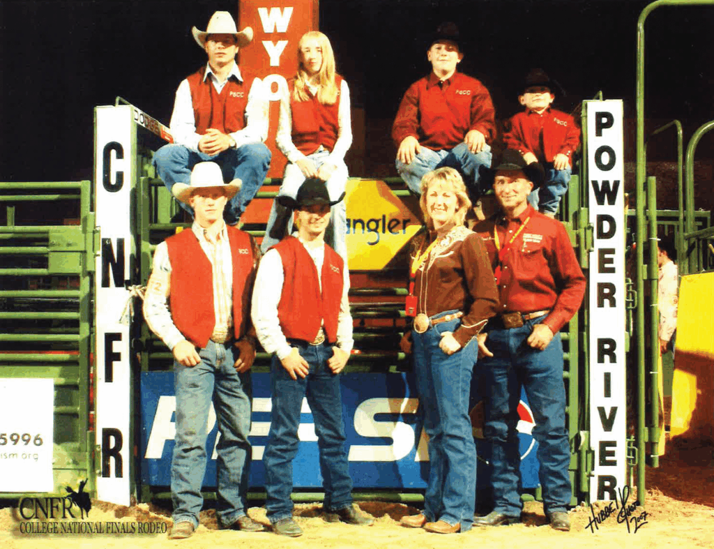 Team photo of the Fort Scott Community College rodeo team at the College National Finals Rodeo. Four are seated on green chutes labeled 'Powder River' and 'CNFR,' including two men in FSCC vests and a man and young boy in maroon shirts. Three men and one woman, most in FSCC vests and western wear, stand on the ground in front beside a man in a maroon western shirt. The group wears cowboy hats, jeans, boots, and belt buckles. The CNFR College National Finals Rodeo logo is visible at the bottom left. The background includes green arena fencing and a yellow chute.
