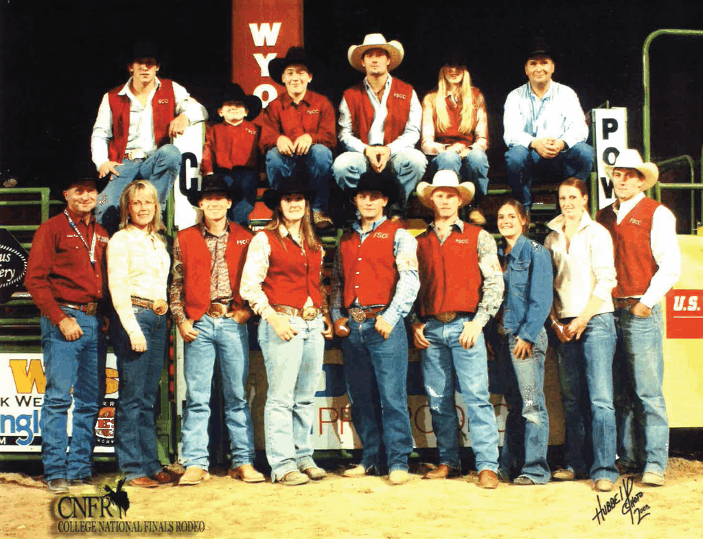 Group photo of the Fort Scott Community College rodeo team at the College National Finals Rodeo. Seven members, including a child and a woman, are seated on green and yellow rodeo chutes labeled 'Powder River,' with nine additional teammates standing in front. Most are wearing FSCC vests, western shirts, jeans, boots, and cowboy hats or belt buckles. The CNFR College National Finals Rodeo logo appears at the bottom left, with green livestock panels and arena advertisements visible in the background on a dirt floor.