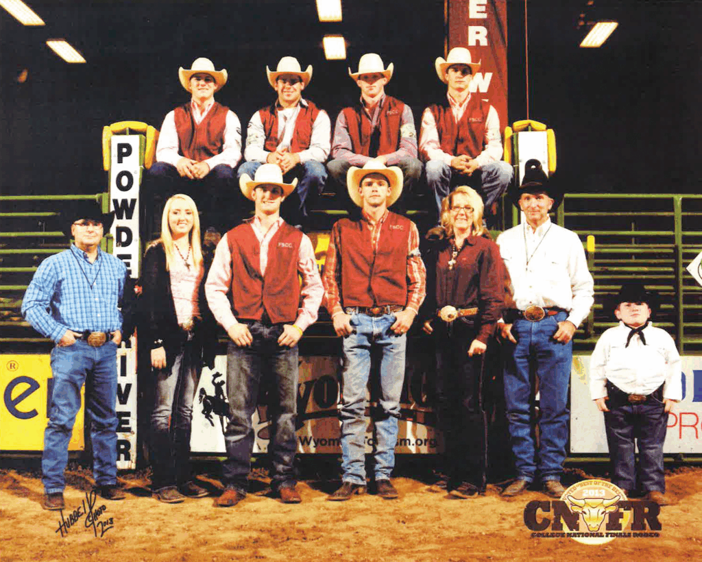Group photograph of the Fort Scott Community College rodeo team taken at an indoor arena during the College National Finals Rodeo 2013. Six young men wearing cowboy hats and FSCC vests are seated on raised yellow and brown chutes labeled 'Powder River,' while five others—men, women, and a child—stand in front. They are dressed in western shirts, jeans, boots, and cowboy hats or belts. A College National Finals Rodeo 2013 logo appears in the bottom right corner, and green railings and arena lights are visible in the background.