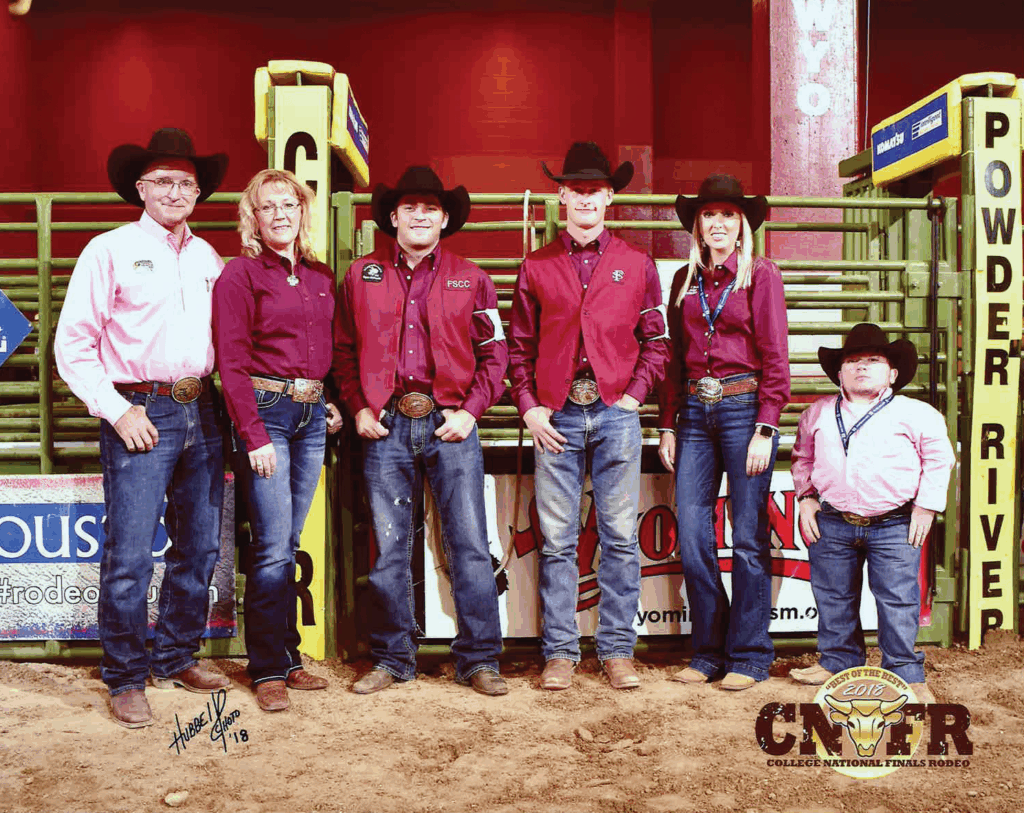 Group photo of the Fort Scott Community College rodeo team taken at the College National Finals Rodeo 2018. Seven members, including a young boy, stand on the dirt arena floor in front of yellow Powder River chutes and a red backdrop. The team wears cowboy hats, maroon FSCC vests or maroon shirts, jeans, boots, and large belt buckles. The CNFR College National Finals Rodeo 2018 logo appears in the bottom right. Green metal railings and arena panels form the background.