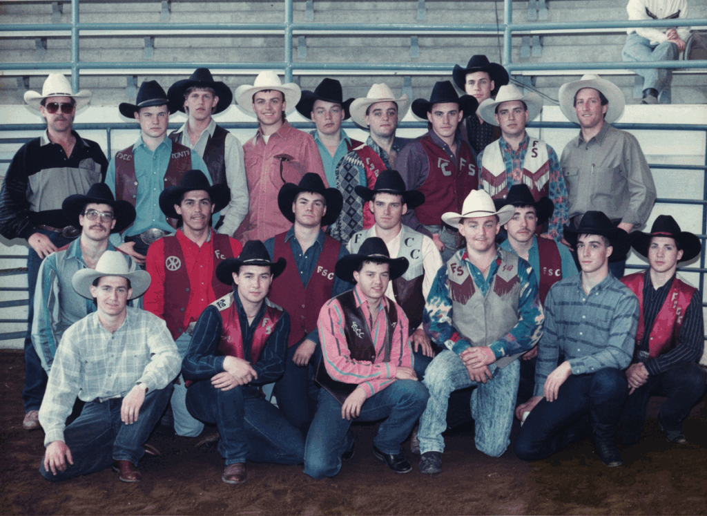 Group photo of a rodeo team with about twenty young men and one adult, all wearing cowboy hats and western shirts or vests, posing together inside an arena with bleachers in the background.